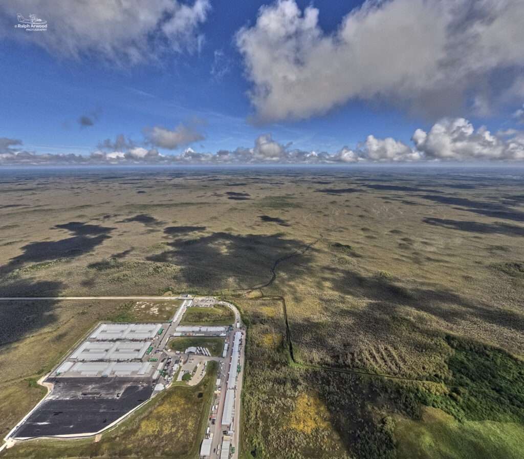 Aerial photo of the Everglades, with portions of the Alligator Alcatraz facility located in the bottom right corner in the photo. A partly cloudy blue sky dominates the upper third.