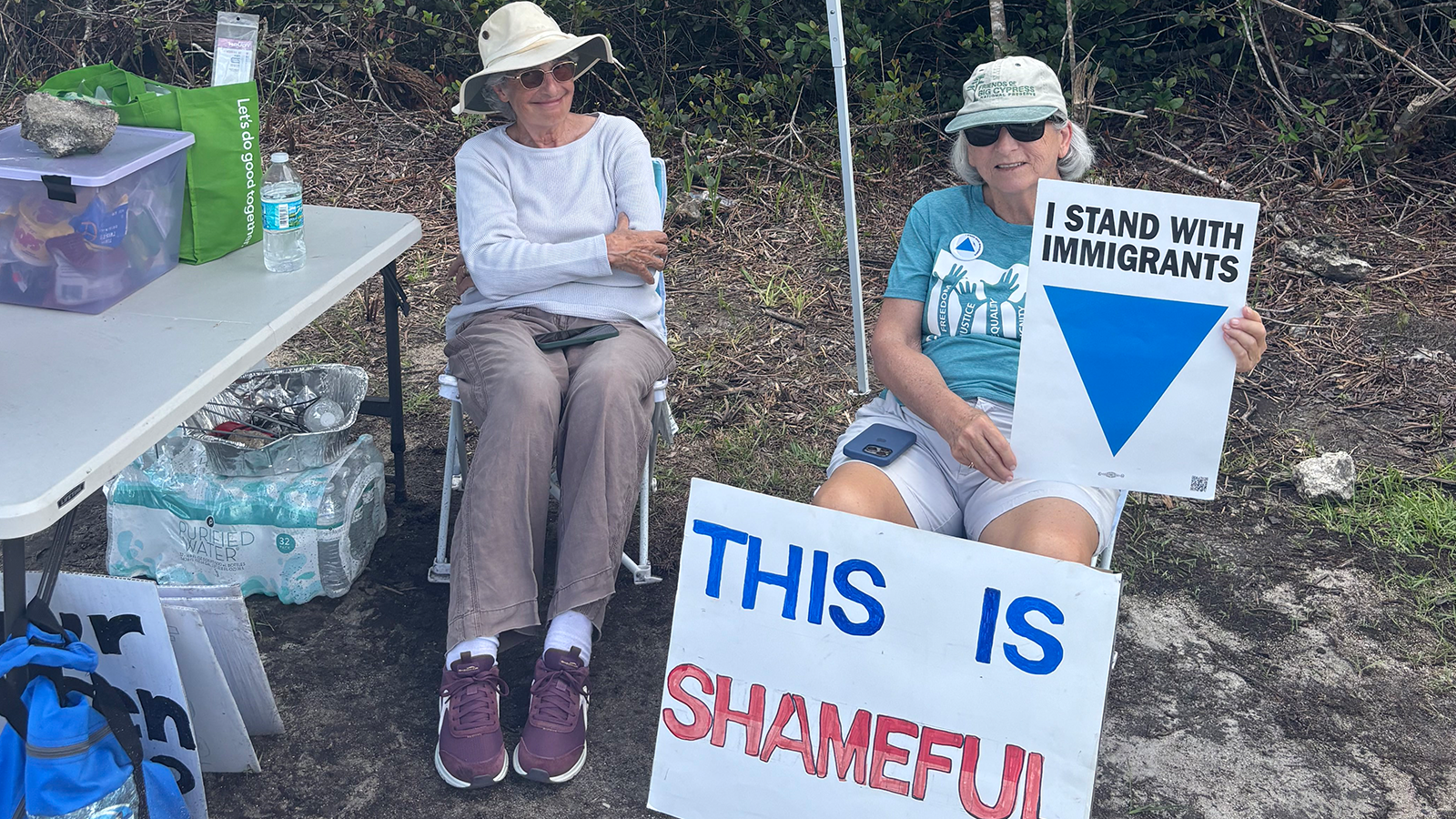 Two women sitting on chairs, one of them has protest signs that read 'I stand with immigrants' and 'this is shameful'