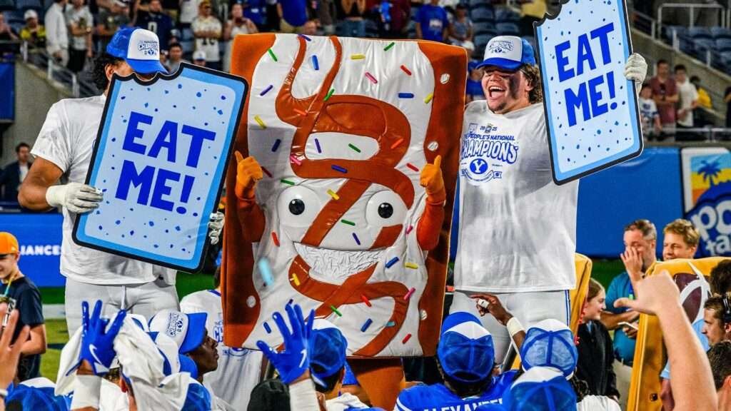 A Pop-Tarts mascot stands next to two football players holding Pop-Tarts-styled signs that say "EAT ME!", surrounded by their teammates.