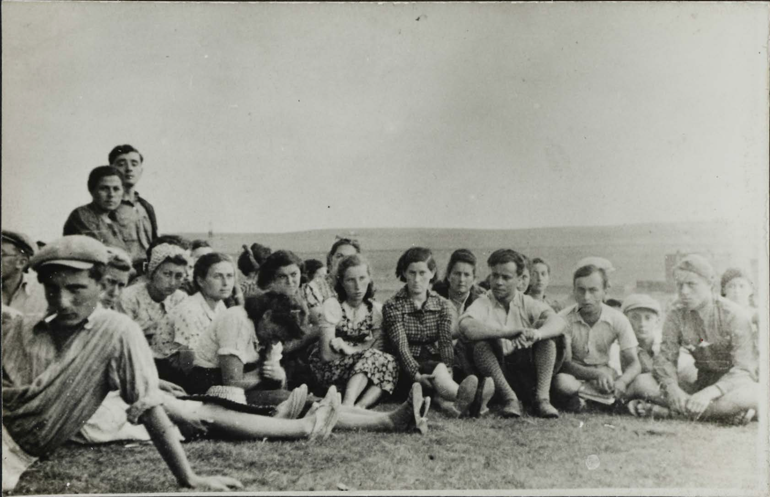 Group of He-Chaluts youth movement members seated on grass outdoors in Będzin, wearing mid-20th-century casual clothing.