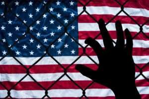 A hand grabs at a chain link fence with an American flag on the other side, symbolizing the struggle to immigrate to America.