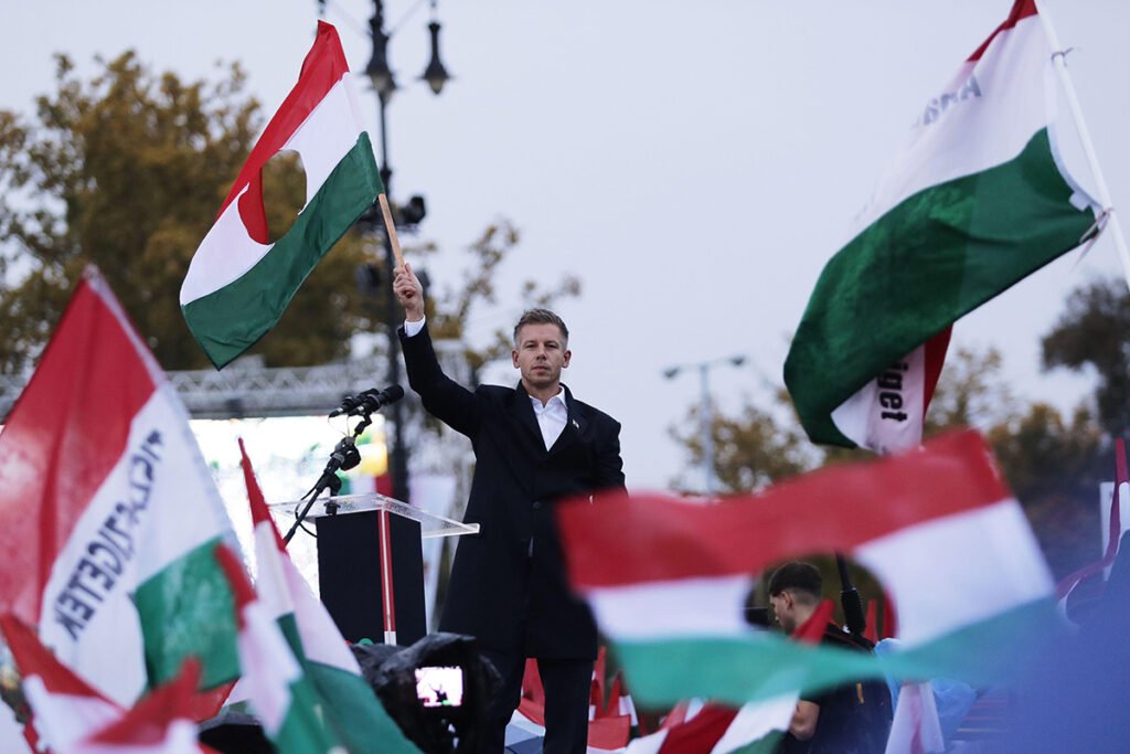 The leader of Hungary’s Tisza party, Peter Magyar, during a demonstration marking the 69th anniversary of the outbreak of Hungary’s 1956 revolution. Photo: AP Photo/Rudolf Karancsi/Alamy

				
				
				
				
				This is an edited version of an article first appeared in the Spring 2026 issue of Index on Censorship, The monster unleashed: How Hungary’s illiberal vision is seducing the Western world, published on 2 April 2026. It has been updated to reflect the outcome of the Hungarian elections on 12 April 2026.
About a year ago, I opened a new email account. It was an encrypted Proton Mail address that journalists are advised to set up early in their careers. I did it rather late, and not because I had suddenly become more reckless with the safety of my sources. On the contrary: for years, there had been so few of them that an extra layer of digital security felt almost unimportant.
Then the messages started arriving. In a few months, the number of emails from people who thought they had something to say reached a level that had to be dealt with. People sent me documents, stories or just tips they thought important. The inbox filled up so quickly that I had to ask myself the question: what had changed?
It is, of course, a fair question to ask how a journalist can work without sources close to power. They cannot. But there is a difference in the kind of sources we have. I am not saying that Hungary does not have its fair share of investigative journalists, but it can be tough for a political journalist working on daily events as I do. Let me explain.
For years I have argued that Hungary is neither a classic dictatorship nor a fully-fledged democracy. The system that Viktor Orbán proudly describes as an “illiberal democracy” has been carefully engineered to reward loyalty and apathy in equal measure. The deal offered by the state is simple and widely understood: you may live comfortably, pursue your private ambitions and be left alone – as long as you do not interfere with ours.
Those who accept this bargain rarely encounter trouble. Those who do not are reminded of the limits of dissent. In theory, freedom of expression and the right to protest are guaranteed by the constitution. In practice, the price of speaking out rises sharply once your voice carries beyond the private sphere.
This is not about heated arguments at family lunches or angry comments left under Facebook posts. It is about teachers who participated in acts of civil disobedience and lost their jobs. Judges whose rulings displeased the government and who subsequently found themselves the subject of orchestrated smear campaigns. University lecturers and civil servants who pointed to structural injustices and were quietly dismissed. It is also about ordinary employees summoned by their managers and warned that their political views, expressed online, were not appreciated.
Silence is survival
In smaller towns and villages, where everyone knows everyone else, the consequences can be even more immediate. Access to public employment schemes is a lifeline for many families, and they often feel that it depends on the goodwill of a mayor aligned with the ruling party. Under such conditions, political neutrality becomes a survival strategy.
The effect of these experiences has been profound. Over the past decade and a half, Hungary has developed a political culture in which self-censorship is not imposed by law but internalised by habit. People do not remain silent because they are forbidden to speak but because they have learned that silence is safer.
For journalists, this climate has had predictable consequences. In a system built on fear, loyalty and informal punishment, ordinary citizens are understandably reluctant to contact the media. Some do, and their courage should not be underestimated. Most, however, choose discretion. Hospital staff, teachers and employees of state-owned companies are far more likely to endure abuses of power than to report them. As a result, Hungarian journalism has long relied less on whistleblowers and citizens than on leaks from within political and economic elites – sources that are themselves increasingly scarce.
This is why the sudden surge of messages mattered. About a year ago, something began to shift. My colleagues and I noticed it almost simultaneously. Emails arrived from people we had never spoken to before. Old landlines started ringing again. In one case, an envelope arrived by post, handwritten, containing detailed suggestions for investigations. Large numbers of Hungarians, many of them previously invisible to the press, were reaching out.
In a media environment where the government does everything it can to restrict access to information, this felt like an unexpected privilege. Independent journalists are routinely excluded from press conferences, denied interviews or simply ignored when they submit questions. At times the situation has bordered on the absurd. There have been moments when we have had to publicly ask our readers to inform us if the prime minister is visiting their town because official channels no longer bother to tell us.
Stepping into the light
So what changed? The short answer is Péter Magyar, the leader of the Tisza Party, appeared. The longer answer is more complicated.
Magyar’s arrival on the political scene was abrupt. A former insider, once a beneficiary of the Orbán system, he had worked as a lawyer at Hungary’s permanent representation to the EU and was married to Judit Varga, then the justice minister. When Varga was forced to resign over a presidential pardon granted to a well-connected figure implicated in a child sexual abuse case, Magyar did not retreat into private life. Instead, he stepped into the spotlight, publicly broke with the ruling Fidesz Party, and founded the Tisza Party.
From last autumn, that party led opinion polls by double digits. [That lead in the opinion polls has since turned into an electroal landslide for Magyar on 12 April.] Few would have predicted such a rapid rise. Fewer still would have predicted the tone Magyar would adopt.
Compared with Orbán’s carefully managed appearances, Magyar is restless, confrontational and informal. He mixes arrogance with humour. He has walked out of a television studio when he disliked the questions.
When a senior Fidesz official shouted at him aggressively in front of cameras, Magyar replied that the man should brush his teeth because of his bad breath. At another event, he offered tea and biscuits to pro-government journalists who had been left waiting in the cold. On Facebook, he comments personally under articles that mention him, often arguing directly with readers.
This behaviour would be unremarkable in some political cultures. In Hungary, it was something new. Magyar appeared unafraid of ridicule, retaliation or scandal. For many, that fearlessness was contagious.
What followed was not simply a surge in public support for a new opposition figure but a subtle loosening of social restraint. People who had long kept their views private began to speak more openly. They attended rallies, shared articles, corrected misinformation and – crucially for journalists – they started to communicate. Tips that would once have been suppressed out of caution now found their way into newsrooms.
This matters because Hungarian journalism has been under sustained pressure since 2010, when Orbán’s Fidesz-KDNP coalition returned to power with a constitutional supermajority. The transformation that followed was immense. Media laws were rewritten, regulation was restructured and oversight bodies were staffed by loyalists. What appeared at first as technical reform laid the groundwork for political control.
The independent media
Ownership proved even more decisive. Once-prominent independent outlets were closed, weakened or absorbed. Népszabadság, for decades a defining voice of Hungarian public life, disappeared overnight. Online portals such as Origo and Index underwent editorial takeovers that ended their independence. In 2018, nearly 500 pro-government outlets were merged into the Central European Press and Media Foundation, a vast conglomerate that now dominates print, broadcast and online media while getting the majority of state advertising.
The result has been a distorted media market. Independent newsrooms operate with fewer resources, shrinking staff numbers and limited reach. Access to official information is restricted, freedom-of-information requests are delayed or ignored, and journalists are routinely excluded from government press conferences. Critical reporters are portrayed in pro-government outlets as foreign agents or enemies of the nation, and some have been put under surveillance using Pegasus spyware – a revelation that further deepened the sense of vulnerability.
But independent journalism has not vanished. It has adapted. Subscription models, reader donations and new digital platforms have allowed a fragile ecosystem to survive. From inside these newsrooms, the past 15 years have felt like a permanent state of emergency.
Another Fidesz victory would have almost certainly confirmed the effectiveness of the government’s media strategy. With public broadcasting firmly under its control and regional media largely aligned, the ruling party faced little resistance in shaping the narrative on issues ranging from domestic opposition to the war in Ukraine.
The financial imbalance is staggering. In the first six months of 2025 alone, Hungary’s public broadcaster received roughly 80 billion Hungarian forints (0 million) in state funding. There was little indication that this flow would slow.
There were, however, darker possibilities. A draft law on “transparency in public life”, periodically revived, would allow the state to blacklist or financially cripple outlets receiving foreign funding. For some independent organisations, that would mean extinction. The closure of Radio Free Europe’s Hungarian service last year and the transfer of the most popular tabloid paper, Blikk, to a pro-government publisher were warnings that the process of taking over the media sphere is not finished.
A time of transition?
Tisza made opposition to this media system a central part of its platform – unsurprisingly, given that Magyar has been one of its main targets. Billboards across the country depicted him as a puppet of Brussels. Pro-government outlets circulate false stories about his party’s policies, including fabricated claims about tax reforms that courts have since ruled to be untrue. Even the prime minister has joined in, sharing AI-generated videos attacking his rival.
In early February, Tisza published its official programme, devoting an entire chapter to public media and access to information. The promises are ambitious: a new media law, restored editorial independence, balanced reporting, transparent standards and the rebuilding of a rural correspondent network. Most controversially, Magyar has pledged to suspend public media news services immediately upon taking power, restarting them only once conditions for impartial reporting have been created.
Whether such promises are realistic now that Magyar has won is another matter. Poland’s recent experience suggests that dismantling a captured public broadcaster without violating rule-of-law norms is extraordinarily difficult.
Hungary’s situation will also be closely monitored by the EU, particularly given the billions of euros in frozen funds tied to democratic safeguards.
A radical overhaul would also involve dismissing large numbers of employees, a move fraught with political risk, and finding replacements in a media landscape where independent journalists number only in the hundreds.
There is also a generational dimension to consider. Many people working in Hungarian journalism today have no memory of a freer media environment. Some of my colleagues were still in school during Orbán’s first years in power. They have never experienced a government that treats the press as a partner rather than an enemy, that invites critical journalists to press conferences, or that answers questions without obstruction.
A transition, if it comes, will not be instantaneous. The new political leadership will have to relearn a basic democratic lesson: the role of the press is not loyalty but scrutiny. Society, too, will have to relearn how to trust journalism. That has been systematically eroded. According to the Reuters Institute’s 2024 Digital News Report, Hungary has the lowest level of trust in news media in Europe. Just 23% of respondents say they trust most news most of the time. Rebuilding that confidence will take years.
And yet the messages keep coming. In that encrypted inbox, amid the noise and uncertainty, there is a fragile sense that fear has loosened its grip. People are testing the limits of speech again, cautiously.
For a journalist in Hungary today, that may be the most hopeful development of all.

			
			
					
				
				
				
				READ MORE