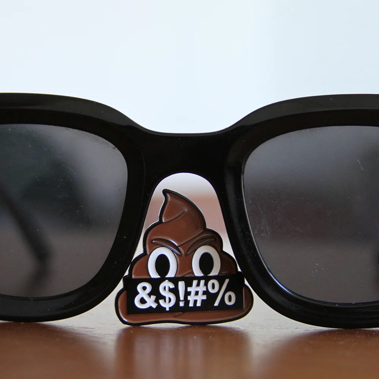 An enamel pin clipped to the nose bridge of black-framed sunglasses resting on a wooden surface. The pin depicts a scowling poop emoji holding a black banner reading "&$!#%".