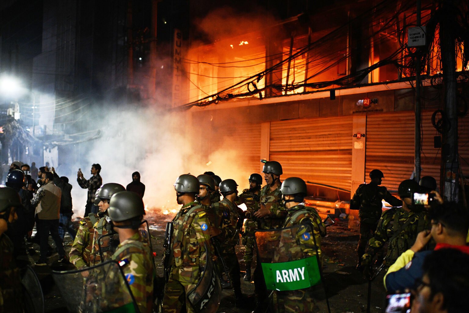 The Bangladeshi army stands guard at the Prothom Alo daily newspaper offices which were set ablaze during protests. Photo: AP Photo/Mahmud Hossain Opu/Alamy
This article first appeared in the Spring 2026 issue of Index on Censorship, The monster unleashed: How Hungary’s illiberal vision is seducing the Western world published on 2 April 2026.
Smoke rose from two buildings late in the Dhaka night, thick and bitter, flames leaping through the shattered windows and gutting newsrooms. Outside, on Kazi Nazrul Islam Avenue and inbang the heart of Karwan Bazar on the night of 18 December 2025, hundreds of people surged forward, chanting, jeering and hurling stones at the offices of Bangladesh’s premier dailies – the Bengali Prothom Alo (First Light) and the English-language Daily Star. Representing the liberal, secular voice of an increasingly divided Bangladesh, editors and journalists at the two newspapers have faced legal attacks from the government and threats of violence. They have also had threats from sections of the general public.
By nightfall, the glass frontage of Prothom Alo’s offices had been smashed. Inside, smoke spread rapidly through the newsroom, curling around desks where reporters had been editing copy only hours earlier. Journalists and staff scrambled for exits as the fire took hold on the lower floors of the building. Across the city, at The Daily Star’s headquarters, a similar scene was unfolding: stones hurled through windows, vehicles torched, entrances blocked and journalists trapped inside as smoke filled stairwells. Videos posted online showed flames licking at the building’s interior as staff shouted for help from the upper floors.
From a neighbouring high-rise, senior Prothom Alo reporter Galib Ashraf watched helplessly as the conflagration gutted the newsroom that had been his professional home for years. “This wasn’t just a building burning,” he told journalists later. “It was our history going up in smoke.” The acrid smell of burning paper was mixed with fear as glass crunched underfoot and sirens wailed in the distance.
For journalists inside the buildings, the experience was visceral. For those watching from outside – fellow reporters, photographers, passers-by – the message was unmistakable. In Bangladesh, even the largest and most established newsrooms were vulnerable to attack.
A month later, when Index visited the charred remains of the two offices, a yellow tape surrounded the building, marking the scene of a crime. Veteran journalist Matiur Rahman, editor of Prothom Alo, forced a smile. “We reached out to everyone that night for help,” he said.
On the night of the fire, several journalists at The Daily Star found themselves trapped in their offices. The only escape route was upwards, to the rooftop. One reporter, Zyma Islam, posted on Facebook from inside, her words chilling in their simplicity: “I can’t breathe anymore … there’s too much smoke … I am inside.” Some reporters feared that they would die. Mahfuz Anam, editor of the Daily Star told Index that if he had been around, he would have been lynched. He, too, tried reaching out to the authorities. Whilst everyone was sympathetic, it took a long time for help to arrive.
The need for a new leader
There was an acute sense of betrayal also hanging in the air when Index visited the offices of the two newspapers. Both Prothom Alo and The Daily Star had argued for a more liberal political order in Bangladesh. In August 2024, the Prime Minister Sheikh Hasina had fled the country when her government agents killed more than 800 student demonstrators. Like many of the country’s educated and middle classes, journalists felt Bangladesh needed a new leader known for probity, someone like the Nobel Laureate Muhammed Yunus. The newspapers supported Yunus, who was known for his pioneering microlending work in Bangladesh, specifically to support indigenous trading women. The fact that he had been persecuted and by Hasina added to his credibility.
So there was considerable enthusiasm when he agreed to become the chief adviser of the interim government, the de facto prime minister until elections were held. But his political choices stunned people. Even while expressing faith in the youth, he blindsided the female student leaders responsible for the uprising that felled Hasina by letting the misogynistic Jamaat-e-Islami party dictate terms. Sheikh Hasina had banned the Jamaat, but following her ousting, the Jamaat had a new lease of life and was going to contest the February elections. Disappointed women leaders of the movement left the newly-formed National Citizen Party that the students had formed. Many felt that Yunus had betrayed their hopes.
On the night of the attacks, the editors and senior journalists, including editors of rival newspapers, made frantic calls to Yunus and his advisers, as well as senior government officials, pleading for help. None came for a long time – both offices had been reduced to burnt-out shells before the fires were brought under control. There was no explanation forthcoming as to why the police did not arrive at the two newspaper offices, in the heart of Dhaka, to stop the mob before the crowds grew in size.
The Daily Star was founded in 1991, two decades after the brutal civil war against Pakistan that led to Bangladesh’s independence – albeit at the cost of hundreds of thousands of lives. Prothom Alo was formed in 1998. The two newspapers have long been recognised as constructive critics of the governments of the day. While both have been accused of pro-India and anti-fundamentalist bias, they are both in fact remarkably independent. Nothing demonstrates this clearer than their dexterous navigation of the tortuous turns of Bangladeshi politics, characterised until recently by the Manichaean divide between the Awami League and the Bangladesh Nationalist Party (BNP) which each took turns holding power since Bangladesh’s independence in 1971.
The night the presses stopped
The December night of the fires, one of the darkest in the history of newspaper publishing in Bangladesh, was not merely a picture of chaos, it was the symbolic decapitation of independent journalism in Bangladesh. The two newspapers were forced to suspend their print editions, Prothom Alo for the first time in nearly three decades and The Daily Star for the first time in its 35-year existence. Martial law, threats of lawsuits and the arrests and disappearances of reporters hadn’t silenced them. But a mob did succeed where others had failed, even if only for one night.
The mobs that converged on Dhaka’s media hubs did not emerge from a vacuum. The proximate cause of the confrontation was the assassination of a student leader, Sharif Osman Hadi, who was the spokesperson for Inquilab Mancha, Platform of the Revolution, which had emerged from the student-led uprising. His killers are still at large. But that anger was quickly and violently redirected at the press. Mobs accused the two newspapers of political bias, branding them “India-backed” and loyal to Hasina.
Human rights and press bodies across the world condemned the attacks, not as isolated incidents but rather as symptoms of a deeper malaise. Irene Khan, the UN Special Rapporteur for freedom of expression and opinion, called the arson attacks ‘deeply alarming,’ expressing her outrage over ‘orchestrated mob violence.’
Bangladesh’s media landscape had been corroded for years by oppressive laws, intimidation and impunity. Whilst the arson attacks were dramatic, they were not anomalous. They were the logical culmination of a long, grinding war on free expression in Bangladesh.
The erosion began with fear. For more than a decade, Bangladesh has stayed in the bottom quartile of global press freedom rankings (in 2025, it ranked 149th out of 180 countries surveyed by Reporters Without Borders). Editors came to understand which stories would invite legal trouble. Reporters learned when not to quote certain sources. Bloggers discovered that a Facebook post could carry the same risks as an investigative exposé. Some were hacked to death, and many fled to safety, seeking asylum abroad.
Digital dissent
A major turning point came in 2018, with the enactment of the Digital Security Act (DSA) – a broad and vaguely worded law ostensibly aimed at combating cybercrime and digital harm. In practice, it became a powerful tool for muzzling dissent. The Act’s provisions criminalised a wide range of speech perceived as “false” or “offensive”, leaving journalists, social-media users and activists vulnerable to long jail terms and heavy fines. Rights groups warned early on that the law could and would be abused to silence critics.
One of the most emblematic cases involved Shamsuzzaman Shams, a reporter for Prothom Alo arrested in 2023 after writing about rising food prices. The Hasina government charged him under the DSA with spreading “false news”. Nearly 3,000 people, including hundreds of journalists, have been charged under the Act since it was passed into law – this in a country whose constitution guarantees freedom of expression.
The DSA’s broad reach is part of a larger pattern, wherein legal mechanisms intended to protect citizens instead serve as implements of fear and silence. In 2020, cartoonist Ahmed Kabir Kishore, for example, spent 10 months in pre-trial detention under the DSA on account of his satirical work, drawing international condemnation for his treatment. The writer Mushtaq Ahmed died in jail in 2021. He had criticised the government’s handling of the pandemic and died of a heart attack, although his supporters and lawyers, including the co-accused, said he had been tortured in jail.
Beyond arrests and lawsuits, the threat of violence hangs like Damocles’ sword over independent voices. One of the starkest and most haunting chapters in Bangladesh’s press freedom story is the disappearance of journalists – most notoriously that of Shafiqul Islam Kajol, a photojournalist and editor who vanished in March 2020. Kajol was last seen leaving his Dhaka home a day after being charged under the DSA with a defamation suit, filed by an Awami League politician. CCTV footage showed unidentified men tampering with his motorcycle before he disappeared. Kajol’s family suspected abduction; rights groups demanded investigations that never yielded closure. Authorities denied he was in custody. When Kajol was eventually found, with his hands and feet tied, near the Indian border, Bangladeshi authorities arrested him for trespassing.
A narrowing conversation
These attacks and deaths are horrifying reminders that journalists and independent thinkers constantly face mortal danger in Bangladesh for the very act of thinking and speaking freely. When journalists fear for their safety and media houses are targets of mob violence, the public conversation narrows. Citizens lose access to independent verification of facts, analysis and accountability reporting, making it easier for misinformation to flourish
In the elections in February 2026, the Bangladesh Nationalist Party (BNP) led by former Prime Minister Khaleda Zia’s son, Tarique Rahman, secured a two-thirds majority in parliamentary seats, with their rivals the Awami League not allowed to stand, and Jamaat performing much less well than people had feared.
While the BNP’s manifesto has spoken of upholding press freedom, in the binary nature of Bangladeshi politics, it might in fact mean that publications suppressed under the Awami League will have greater freedom – while publications that opposed the BNP might find that not much has changed. A newspaper which BNP leader Rahman was involved with was accused of running campaigns against atheist bloggers.
What distinguishes the recent attacks on Prothom Alo and The Daily Star however was not merely their scale, but their symbolism. This was censorship by arson, carried out not by the state directly, but by crowds emboldened by years of official hostility. When governments describe journalists as enemies, traitors or foreign agents, they license others to act accordingly.
When attacks on critics of the government are normalised, the moral fabric of society frays. Bengal is the culture of patrikas, pamphlets penned by intellectuals to defy orthodoxy. Shut them down, and it becomes a lesser Bengal. When voices are silenced or endangered, the very sense of a collective narrative – of what holds a diverse nation together – is weakened.
READ MORE The Bangladeshi army stands guard at the Prothom Alo daily newspaper offices which were set ablaze during protests. Photo: AP Photo/Mahmud Hossain Opu/Alamy
This article first appeared in the Spring 2026 issue of Index on Censorship, The monster unleashed: How Hungary’s illiberal vision is seducing the Western world published on 2 April 2026.
Smoke rose from two buildings late in the Dhaka night, thick and bitter, flames leaping through the shattered windows and gutting newsrooms. Outside, on Kazi Nazrul Islam Avenue and inbang the heart of Karwan Bazar on the night of 18 December 2025, hundreds of people surged forward, chanting, jeering and hurling stones at the offices of Bangladesh’s premier dailies – the Bengali Prothom Alo (First Light) and the English-language Daily Star. Representing the liberal, secular voice of an increasingly divided Bangladesh, editors and journalists at the two newspapers have faced legal attacks from the government and threats of violence. They have also had threats from sections of the general public.
By nightfall, the glass frontage of Prothom Alo’s offices had been smashed. Inside, smoke spread rapidly through the newsroom, curling around desks where reporters had been editing copy only hours earlier. Journalists and staff scrambled for exits as the fire took hold on the lower floors of the building. Across the city, at The Daily Star’s headquarters, a similar scene was unfolding: stones hurled through windows, vehicles torched, entrances blocked and journalists trapped inside as smoke filled stairwells. Videos posted online showed flames licking at the building’s interior as staff shouted for help from the upper floors.
From a neighbouring high-rise, senior Prothom Alo reporter Galib Ashraf watched helplessly as the conflagration gutted the newsroom that had been his professional home for years. “This wasn’t just a building burning,” he told journalists later. “It was our history going up in smoke.” The acrid smell of burning paper was mixed with fear as glass crunched underfoot and sirens wailed in the distance.
For journalists inside the buildings, the experience was visceral. For those watching from outside – fellow reporters, photographers, passers-by – the message was unmistakable. In Bangladesh, even the largest and most established newsrooms were vulnerable to attack.
A month later, when Index visited the charred remains of the two offices, a yellow tape surrounded the building, marking the scene of a crime. Veteran journalist Matiur Rahman, editor of Prothom Alo, forced a smile. “We reached out to everyone that night for help,” he said.
On the night of the fire, several journalists at The Daily Star found themselves trapped in their offices. The only escape route was upwards, to the rooftop. One reporter, Zyma Islam, posted on Facebook from inside, her words chilling in their simplicity: “I can’t breathe anymore … there’s too much smoke … I am inside.” Some reporters feared that they would die. Mahfuz Anam, editor of the Daily Star told Index that if he had been around, he would have been lynched. He, too, tried reaching out to the authorities. Whilst everyone was sympathetic, it took a long time for help to arrive.
The need for a new leader
There was an acute sense of betrayal also hanging in the air when Index visited the offices of the two newspapers. Both Prothom Alo and The Daily Star had argued for a more liberal political order in Bangladesh. In August 2024, the Prime Minister Sheikh Hasina had fled the country when her government agents killed more than 800 student demonstrators. Like many of the country’s educated and middle classes, journalists felt Bangladesh needed a new leader known for probity, someone like the Nobel Laureate Muhammed Yunus. The newspapers supported Yunus, who was known for his pioneering microlending work in Bangladesh, specifically to support indigenous trading women. The fact that he had been persecuted and by Hasina added to his credibility.
So there was considerable enthusiasm when he agreed to become the chief adviser of the interim government, the de facto prime minister until elections were held. But his political choices stunned people. Even while expressing faith in the youth, he blindsided the female student leaders responsible for the uprising that felled Hasina by letting the misogynistic Jamaat-e-Islami party dictate terms. Sheikh Hasina had banned the Jamaat, but following her ousting, the Jamaat had a new lease of life and was going to contest the February elections. Disappointed women leaders of the movement left the newly-formed National Citizen Party that the students had formed. Many felt that Yunus had betrayed their hopes.
On the night of the attacks, the editors and senior journalists, including editors of rival newspapers, made frantic calls to Yunus and his advisers, as well as senior government officials, pleading for help. None came for a long time – both offices had been reduced to burnt-out shells before the fires were brought under control. There was no explanation forthcoming as to why the police did not arrive at the two newspaper offices, in the heart of Dhaka, to stop the mob before the crowds grew in size.
The Daily Star was founded in 1991, two decades after the brutal civil war against Pakistan that led to Bangladesh’s independence – albeit at the cost of hundreds of thousands of lives. Prothom Alo was formed in 1998. The two newspapers have long been recognised as constructive critics of the governments of the day. While both have been accused of pro-India and anti-fundamentalist bias, they are both in fact remarkably independent. Nothing demonstrates this clearer than their dexterous navigation of the tortuous turns of Bangladeshi politics, characterised until recently by the Manichaean divide between the Awami League and the Bangladesh Nationalist Party (BNP) which each took turns holding power since Bangladesh’s independence in 1971.
The night the presses stopped
The December night of the fires, one of the darkest in the history of newspaper publishing in Bangladesh, was not merely a picture of chaos, it was the symbolic decapitation of independent journalism in Bangladesh. The two newspapers were forced to suspend their print editions, Prothom Alo for the first time in nearly three decades and The Daily Star for the first time in its 35-year existence. Martial law, threats of lawsuits and the arrests and disappearances of reporters hadn’t silenced them. But a mob did succeed where others had failed, even if only for one night.
The mobs that converged on Dhaka’s media hubs did not emerge from a vacuum. The proximate cause of the confrontation was the assassination of a student leader, Sharif Osman Hadi, who was the spokesperson for Inquilab Mancha, Platform of the Revolution, which had emerged from the student-led uprising. His killers are still at large. But that anger was quickly and violently redirected at the press. Mobs accused the two newspapers of political bias, branding them “India-backed” and loyal to Hasina.
Human rights and press bodies across the world condemned the attacks, not as isolated incidents but rather as symptoms of a deeper malaise. Irene Khan, the UN Special Rapporteur for freedom of expression and opinion, called the arson attacks ‘deeply alarming,’ expressing her outrage over ‘orchestrated mob violence.’
Bangladesh’s media landscape had been corroded for years by oppressive laws, intimidation and impunity. Whilst the arson attacks were dramatic, they were not anomalous. They were the logical culmination of a long, grinding war on free expression in Bangladesh.
The erosion began with fear. For more than a decade, Bangladesh has stayed in the bottom quartile of global press freedom rankings (in 2025, it ranked 149th out of 180 countries surveyed by Reporters Without Borders). Editors came to understand which stories would invite legal trouble. Reporters learned when not to quote certain sources. Bloggers discovered that a Facebook post could carry the same risks as an investigative exposé. Some were hacked to death, and many fled to safety, seeking asylum abroad.
Digital dissent
A major turning point came in 2018, with the enactment of the Digital Security Act (DSA) – a broad and vaguely worded law ostensibly aimed at combating cybercrime and digital harm. In practice, it became a powerful tool for muzzling dissent. The Act’s provisions criminalised a wide range of speech perceived as “false” or “offensive”, leaving journalists, social-media users and activists vulnerable to long jail terms and heavy fines. Rights groups warned early on that the law could and would be abused to silence critics.
One of the most emblematic cases involved Shamsuzzaman Shams, a reporter for Prothom Alo arrested in 2023 after writing about rising food prices. The Hasina government charged him under the DSA with spreading “false news”. Nearly 3,000 people, including hundreds of journalists, have been charged under the Act since it was passed into law – this in a country whose constitution guarantees freedom of expression.
The DSA’s broad reach is part of a larger pattern, wherein legal mechanisms intended to protect citizens instead serve as implements of fear and silence. In 2020, cartoonist Ahmed Kabir Kishore, for example, spent 10 months in pre-trial detention under the DSA on account of his satirical work, drawing international condemnation for his treatment. The writer Mushtaq Ahmed died in jail in 2021. He had criticised the government’s handling of the pandemic and died of a heart attack, although his supporters and lawyers, including the co-accused, said he had been tortured in jail.
Beyond arrests and lawsuits, the threat of violence hangs like Damocles’ sword over independent voices. One of the starkest and most haunting chapters in Bangladesh’s press freedom story is the disappearance of journalists – most notoriously that of Shafiqul Islam Kajol, a photojournalist and editor who vanished in March 2020. Kajol was last seen leaving his Dhaka home a day after being charged under the DSA with a defamation suit, filed by an Awami League politician. CCTV footage showed unidentified men tampering with his motorcycle before he disappeared. Kajol’s family suspected abduction; rights groups demanded investigations that never yielded closure. Authorities denied he was in custody. When Kajol was eventually found, with his hands and feet tied, near the Indian border, Bangladeshi authorities arrested him for trespassing.
A narrowing conversation
These attacks and deaths are horrifying reminders that journalists and independent thinkers constantly face mortal danger in Bangladesh for the very act of thinking and speaking freely. When journalists fear for their safety and media houses are targets of mob violence, the public conversation narrows. Citizens lose access to independent verification of facts, analysis and accountability reporting, making it easier for misinformation to flourish
In the elections in February 2026, the Bangladesh Nationalist Party (BNP) led by former Prime Minister Khaleda Zia’s son, Tarique Rahman, secured a two-thirds majority in parliamentary seats, with their rivals the Awami League not allowed to stand, and Jamaat performing much less well than people had feared.
While the BNP’s manifesto has spoken of upholding press freedom, in the binary nature of Bangladeshi politics, it might in fact mean that publications suppressed under the Awami League will have greater freedom – while publications that opposed the BNP might find that not much has changed. A newspaper which BNP leader Rahman was involved with was accused of running campaigns against atheist bloggers.
What distinguishes the recent attacks on Prothom Alo and The Daily Star however was not merely their scale, but their symbolism. This was censorship by arson, carried out not by the state directly, but by crowds emboldened by years of official hostility. When governments describe journalists as enemies, traitors or foreign agents, they license others to act accordingly.
When attacks on critics of the government are normalised, the moral fabric of society frays. Bengal is the culture of patrikas, pamphlets penned by intellectuals to defy orthodoxy. Shut them down, and it becomes a lesser Bengal. When voices are silenced or endangered, the very sense of a collective narrative – of what holds a diverse nation together – is weakened.
READ MORE