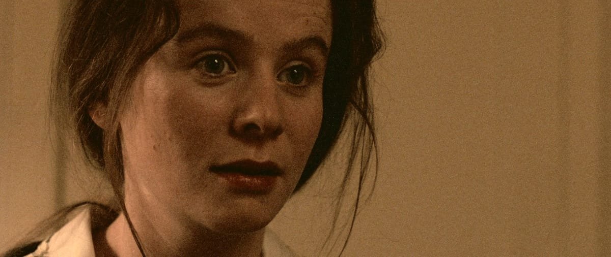 Young woman with dark hair tied back and a white blouse looks ahead in a warmly lit close-up.