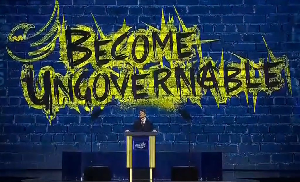 Ross Ulbricht in a suit stands at a podium in front of a blue brick wall featuring a large yellow splash with the words "BECOME UNGOVERNABLE" written in black.
